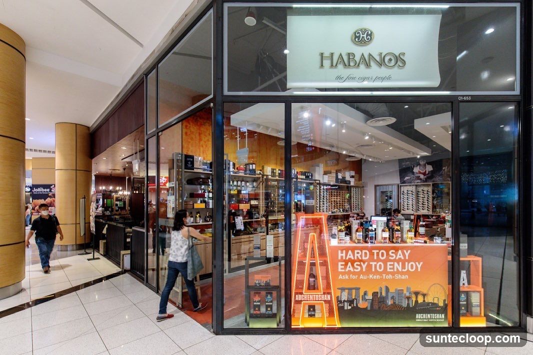 The exterior of a cigar shop with large glass windows displaying various cigar boxes, bottles, and promotional materials. The shop's name, "HABANOS", is prominently displayed above the entrance. A large orange display shaped like the letter 'A' with the brand name "AUCHENTOSHAN" is visible inside.