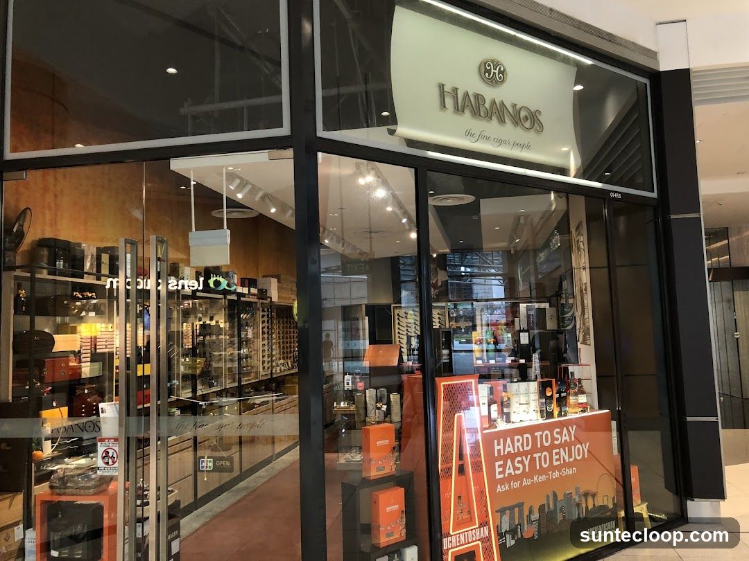 The exterior of a cigar shop with the name "Habanos" displayed prominently above the entrance. The shop window displays various cigar boxes, bottles of liquor, and promotional materials. A sign inside the window reads "HARD TO SAY EASY TO ENJOY Ask for Au-Ken-Toh-Shan".