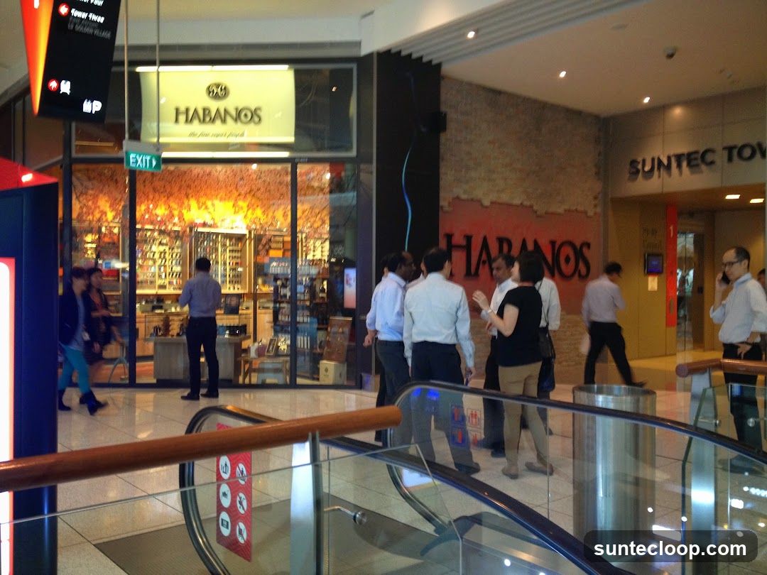 A storefront for a business named Habanos, with a large illuminated sign above the entrance. The store interior is visible through the glass windows, displaying various items on shelves and counters. Several people are standing outside the store and near an escalator.