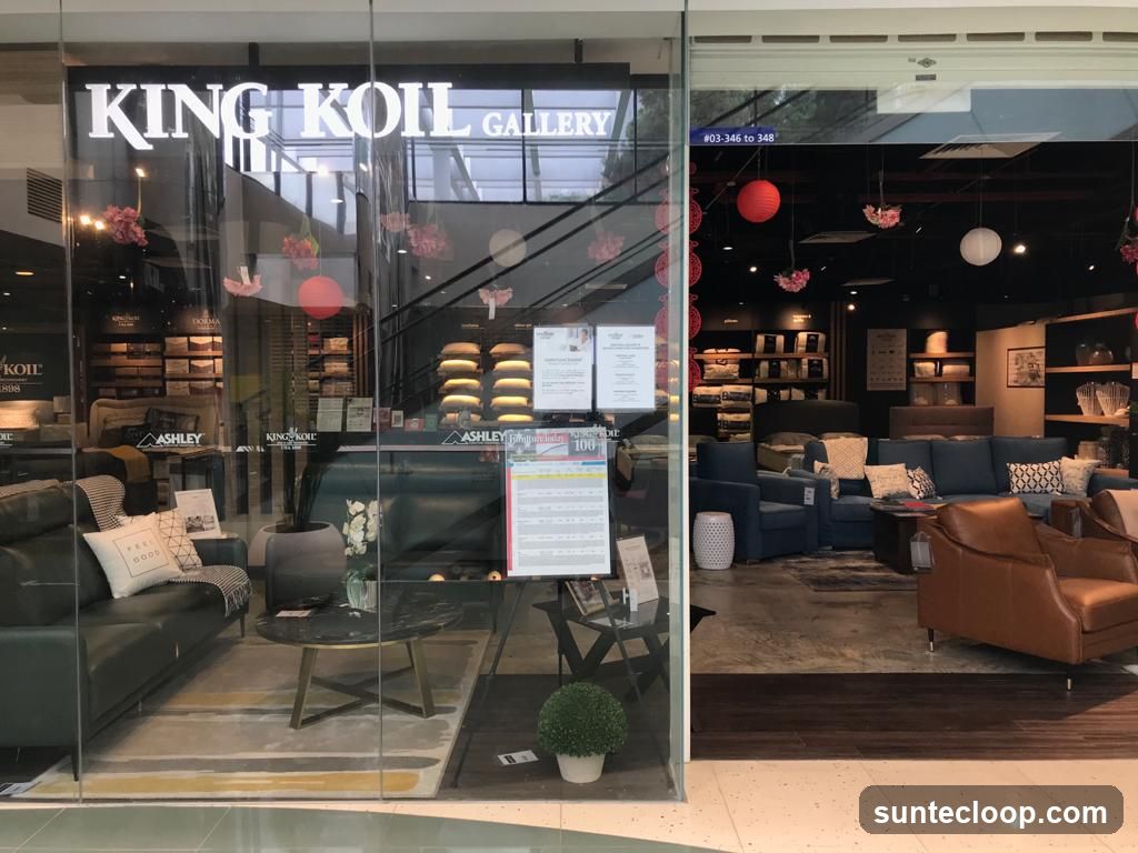 The storefront of King Koil Gallery, a furniture store, is visible through a large glass window. Inside, various sofas, armchairs, beds, and display shelves filled with bedding and pillows are arranged. Decorative elements like red and white lanterns and floral accents are hung from the ceiling. A price list or promotional flyer is displayed on an easel near the entrance.