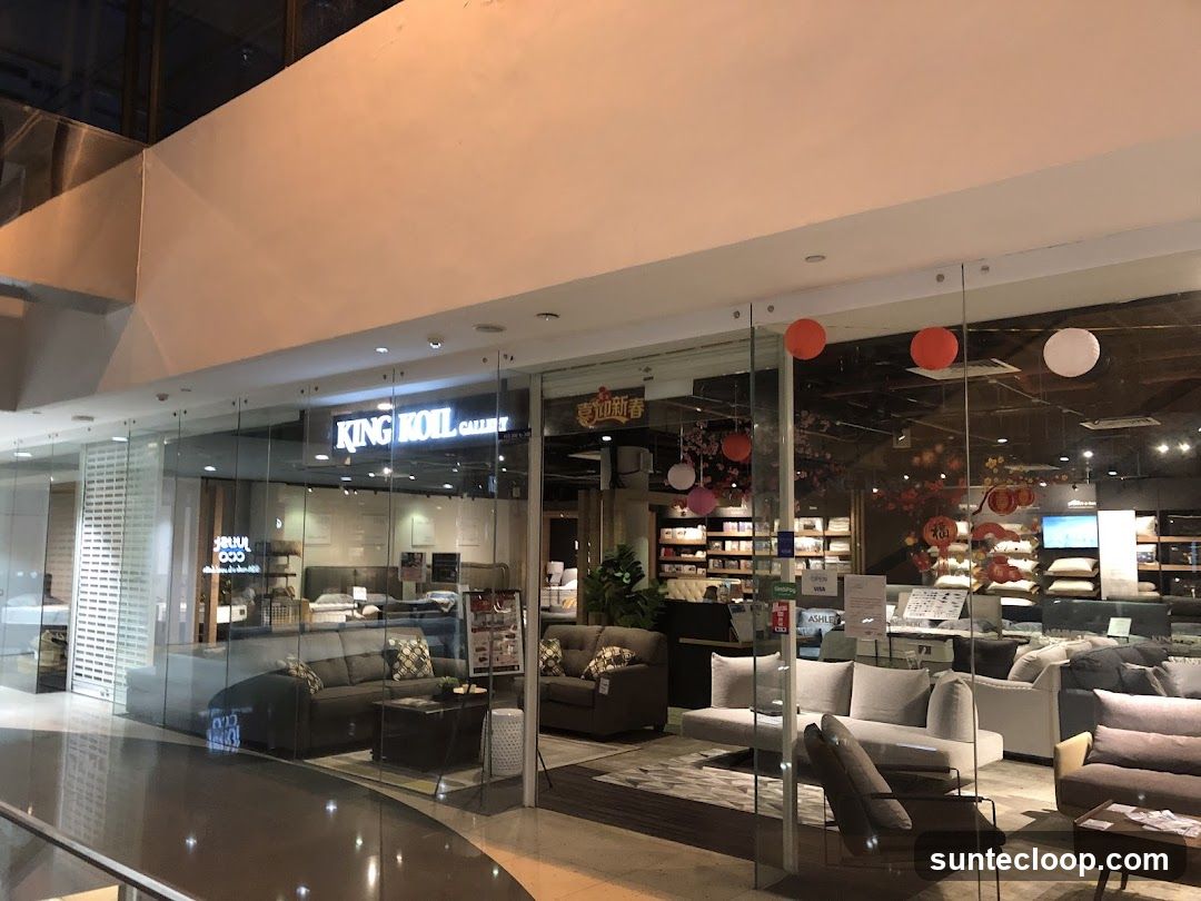 The storefront of King Koil Gallery, a furniture store, is visible through large glass windows. Inside, various sofas, beds, and mattresses are displayed. Decorative lanterns and Chinese New Year decorations are hung inside the store.