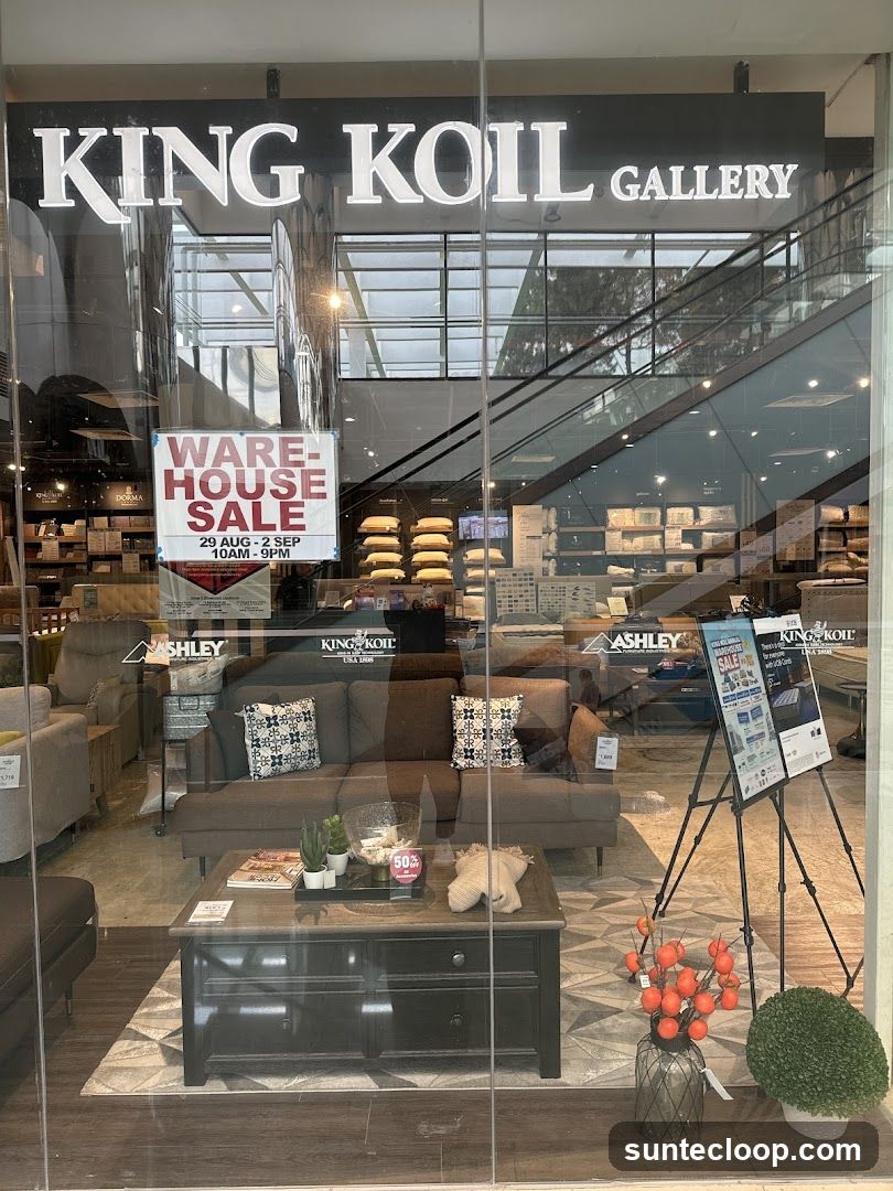 The storefront of King Koil Gallery, a furniture store, is visible through a glass window. The store's name is prominently displayed in large white letters above the entrance. Inside, various furniture items such as sofas, beds, and mattresses are displayed. A "Warehouse Sale" sign is visible on the window, advertising a sale from August 29th to September 2nd, from 10 AM to 9 PM. A coffee table with decorative items is placed in the foreground.