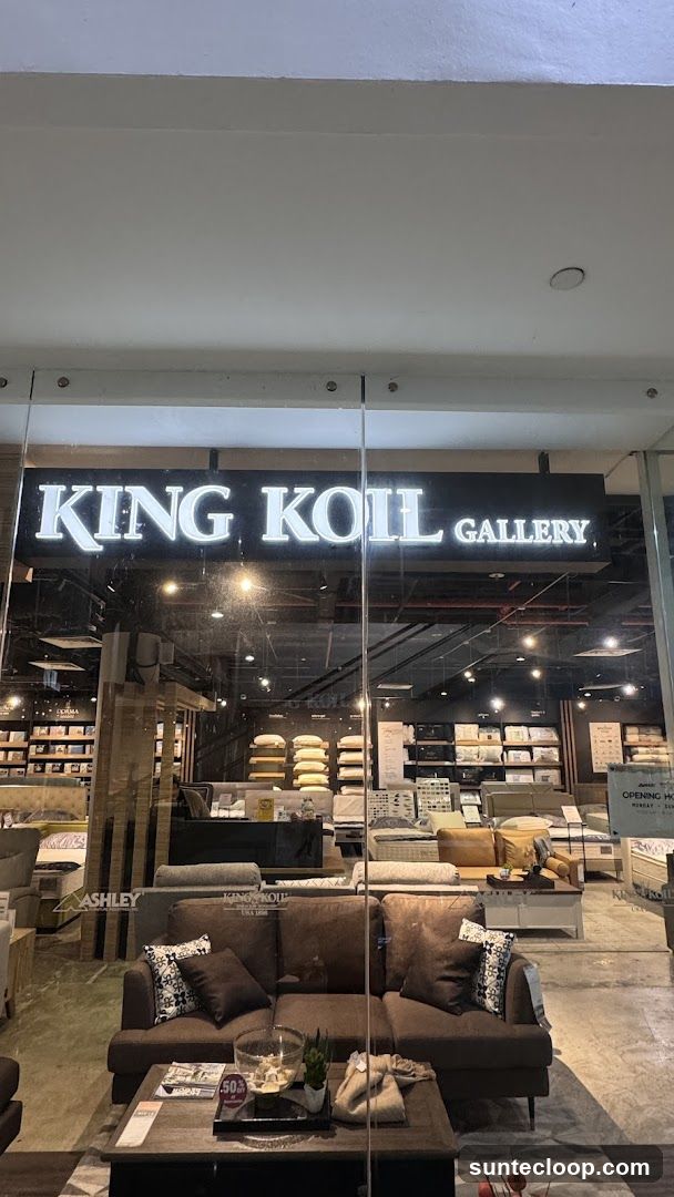 The exterior of a furniture store called King Koil Gallery. The storefront features a large glass window with the store's name illuminated in large white letters above. Inside, various sofas, beds, and mattresses are displayed. A brown sectional sofa is prominently placed in the foreground, with a coffee table in front of it.