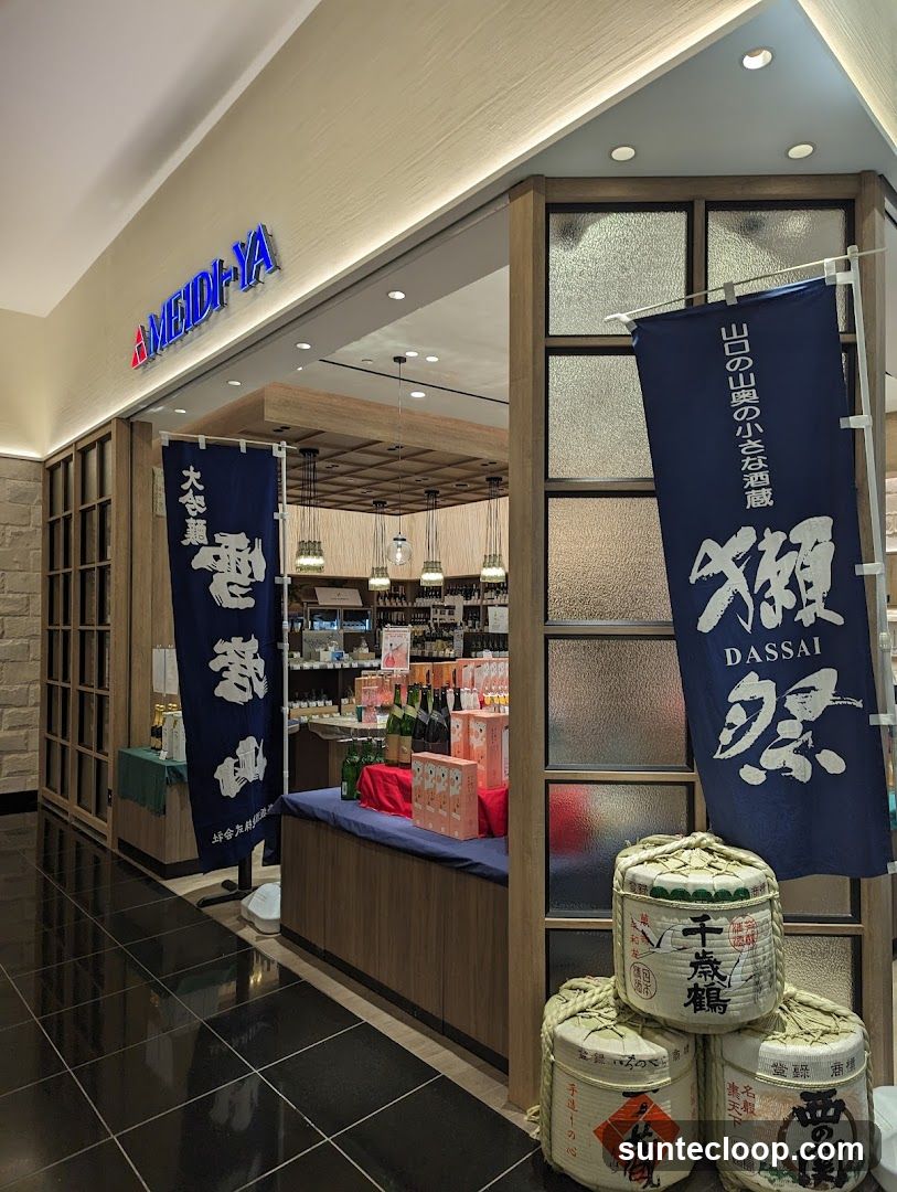 The exterior of a store with the name MEIDI-YA visible above the entrance. Blue banners with Japanese text and images of sake barrels are displayed outside. The store interior, stocked with bottles and boxes, is visible through the open entrance.