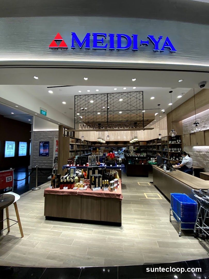 The exterior of a Meidi-Ya store, a wine and liquor shop. The store's name is prominently displayed in blue lettering with a red triangular logo above the entrance. Inside, shelves are stocked with various bottles, and a display table in the foreground showcases more products. A cashier is visible at the counter.