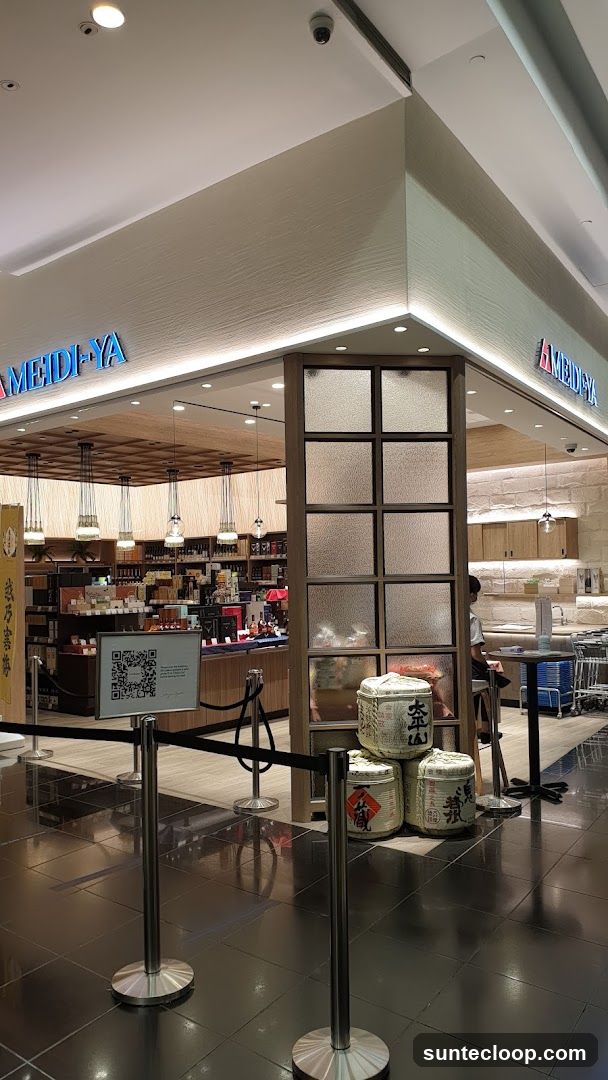The exterior of a Meidi-Ya store with its name displayed in blue neon letters above the entrance. The store features a modern design with wooden accents and frosted glass panels. Inside, shelves are stocked with various products, and decorative sake barrels are displayed near the entrance. A QR code is visible on a stand.