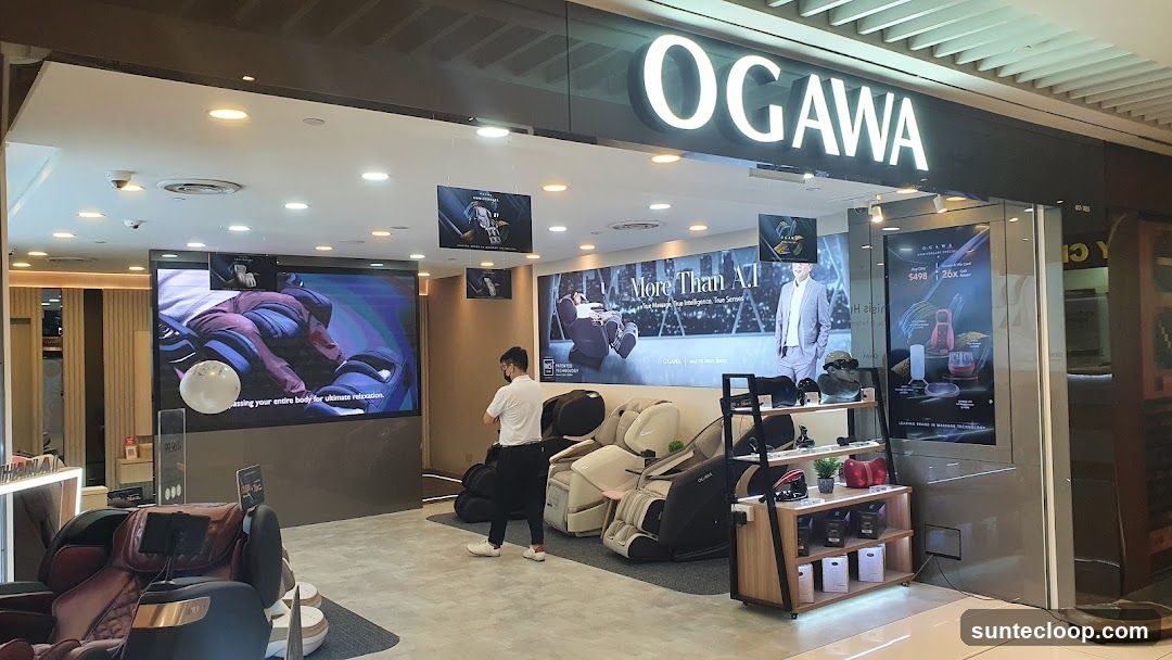 The exterior of an OGAWA store, featuring a large illuminated sign with the brand name. Inside, massage chairs are displayed, along with digital screens showcasing advertisements and product information. A staff member is visible attending to the display.