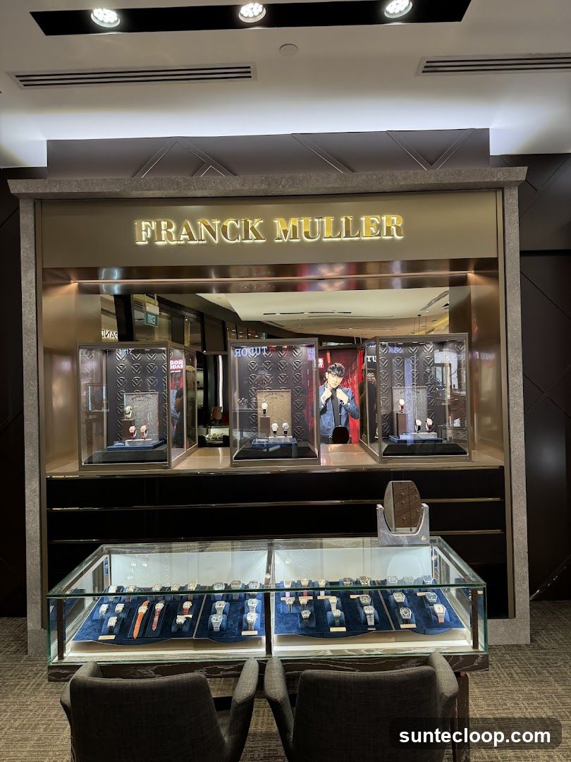 A Franck Muller watch store with illuminated signage above a display of watches. The store features glass display cases showcasing various timepieces, with seating in the foreground. A person is visible behind one of the display cases.