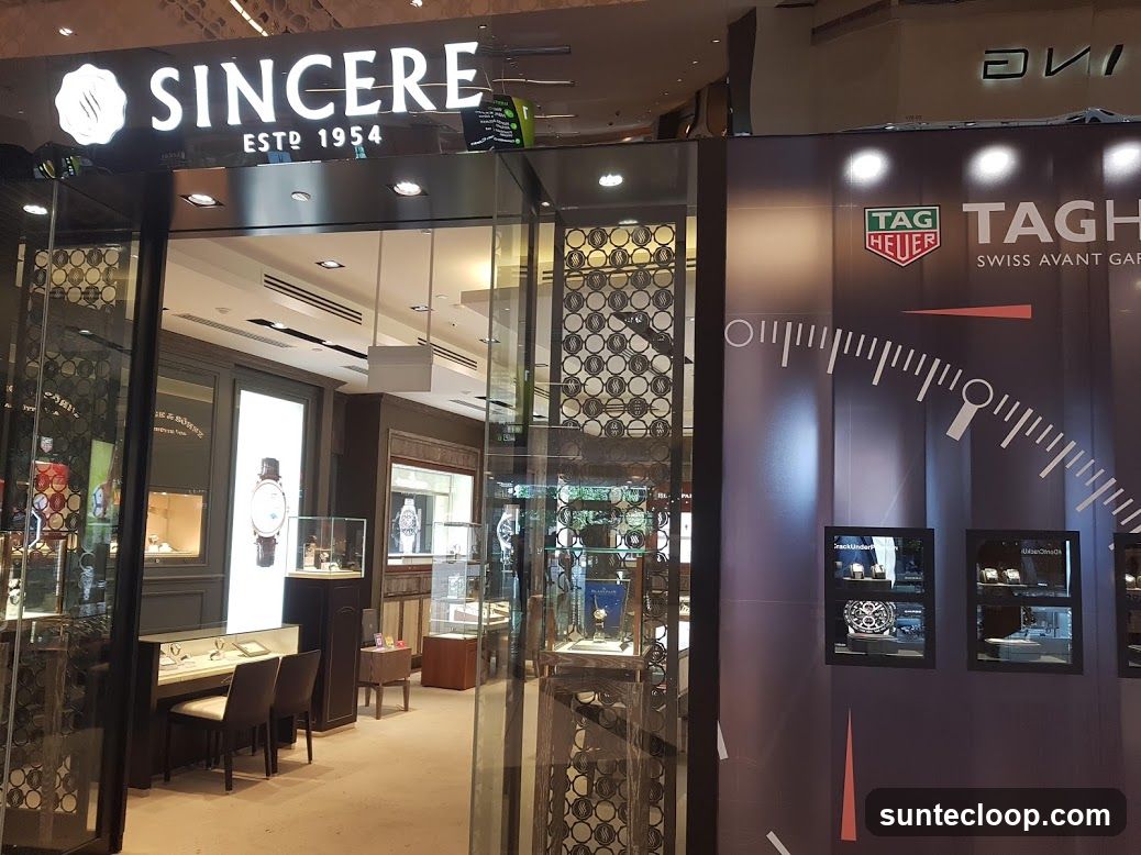 The exterior of a watch store with the name "SINCERE ESTD 1954" prominently displayed above the entrance. The store features large glass windows showcasing various watches and display cases. To the right, there is a large advertisement for TAG Heuer, featuring a graphic of a watch dial and the slogan "SWISS AVANT GARDE".