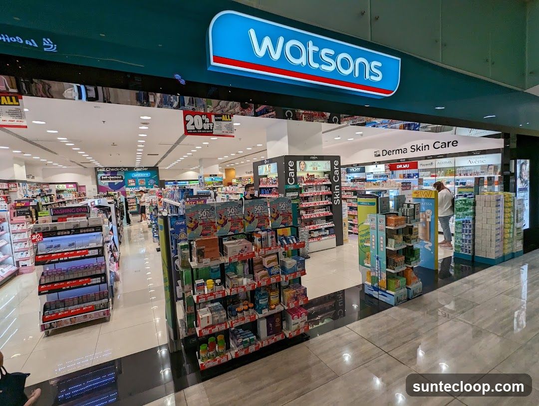 The exterior of a Watsons store, a health and beauty retailer. The prominent Watsons logo is visible above the entrance. Inside, shelves are stocked with various health and beauty products, and promotional displays are set up. A sign indicates "Derma Skin Care" and "Self Check-Out" and "Cashier" stations are visible.