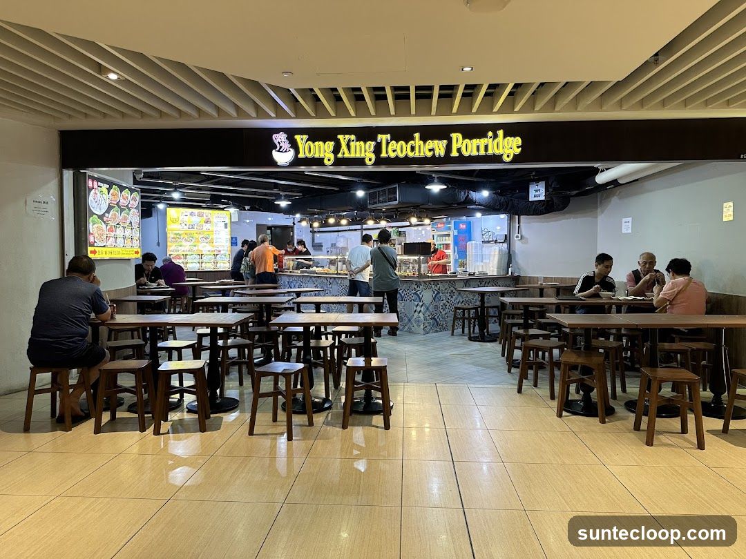 The exterior of a food stall named Yong King Teochew Porridge. The stall has a prominent yellow sign with the name of the establishment. Inside, there are food displays and staff preparing food. Several tables and chairs are set up in front of the stall for customers. Some customers are seated and eating. There are also menu boards displayed on the wall.