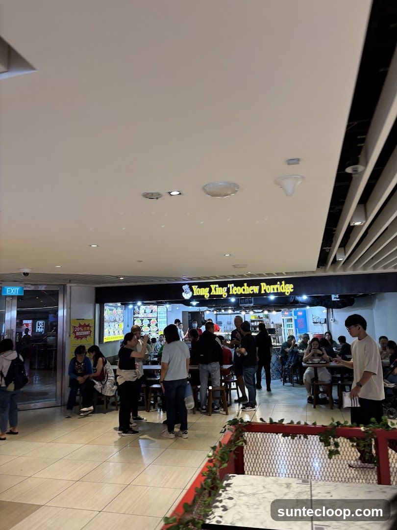A busy food stall with customers seated at tables and standing in line. The stall has a prominent sign that reads "Yong Xing Teochew Porridge." There are menu boards displayed behind the counter, showing various food items. A sign on the left indicates a "20% discount."