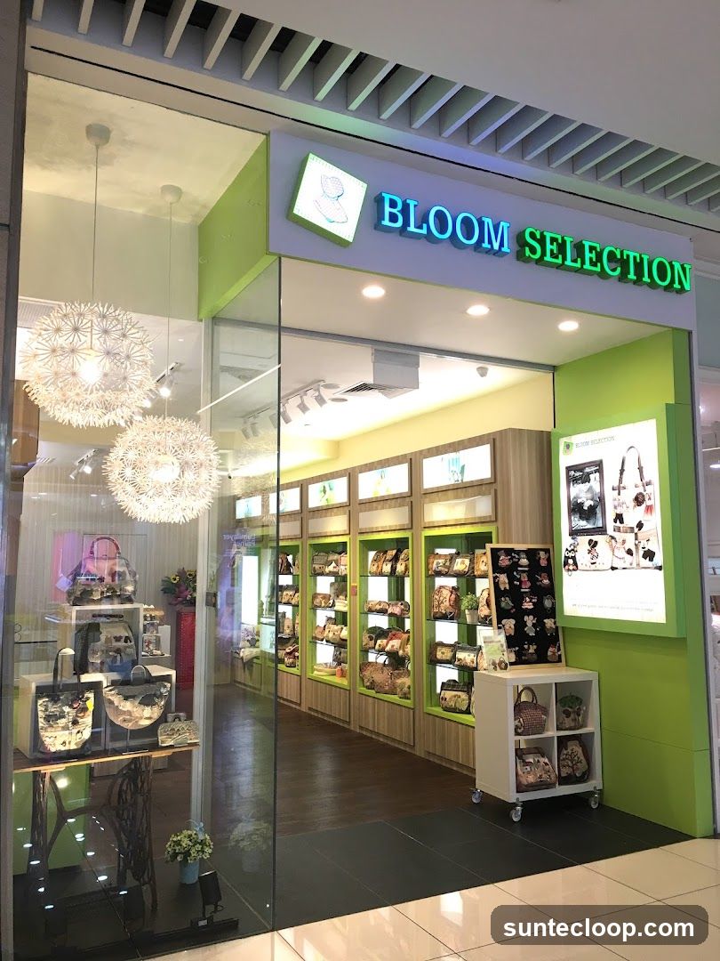 The exterior of a retail store called Bloom Selection, featuring a bright green and white facade. The store's name is displayed in illuminated blue and green lettering above the entrance. Through the glass windows, shelves are visible, stocked with various bags and accessories. Decorative pendant lights hang inside the store.