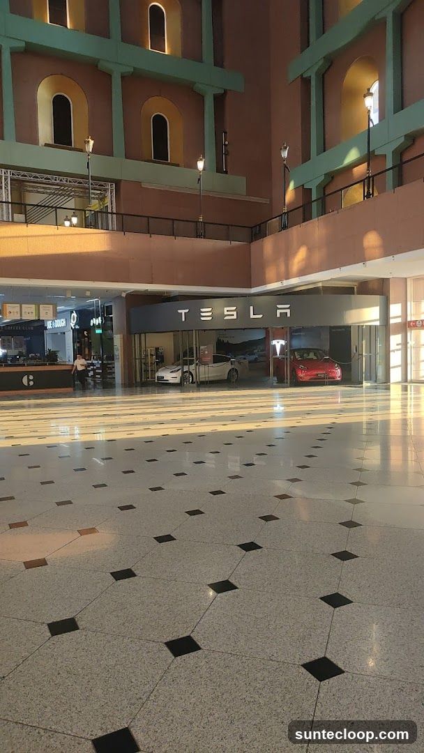 A Tesla showroom is visible with two cars on display, a white one and a red one. The Tesla logo is prominently displayed above the entrance. The showroom appears to be located within a larger building with a distinctive architectural style, featuring arched windows and decorative columns. The floor is tiled with a pattern of light and dark squares.
