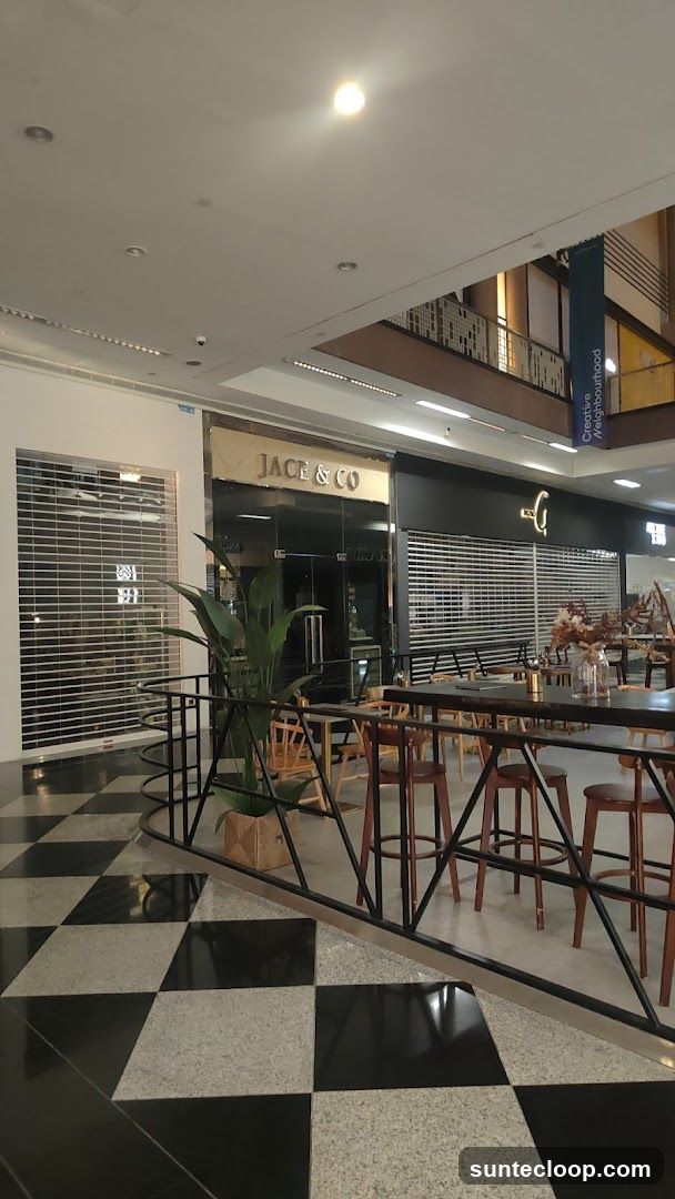 The exterior of a store named Jace & Co. is visible in a shopping mall. The store has a dark facade with a gold sign above the entrance. The entrance features glass doors and windows. To the left of Jace & Co. is another store with its security gate down. In front of the stores, there is a seating area with tables and chairs, and a large potted plant. The floor has a black and white checkered pattern.