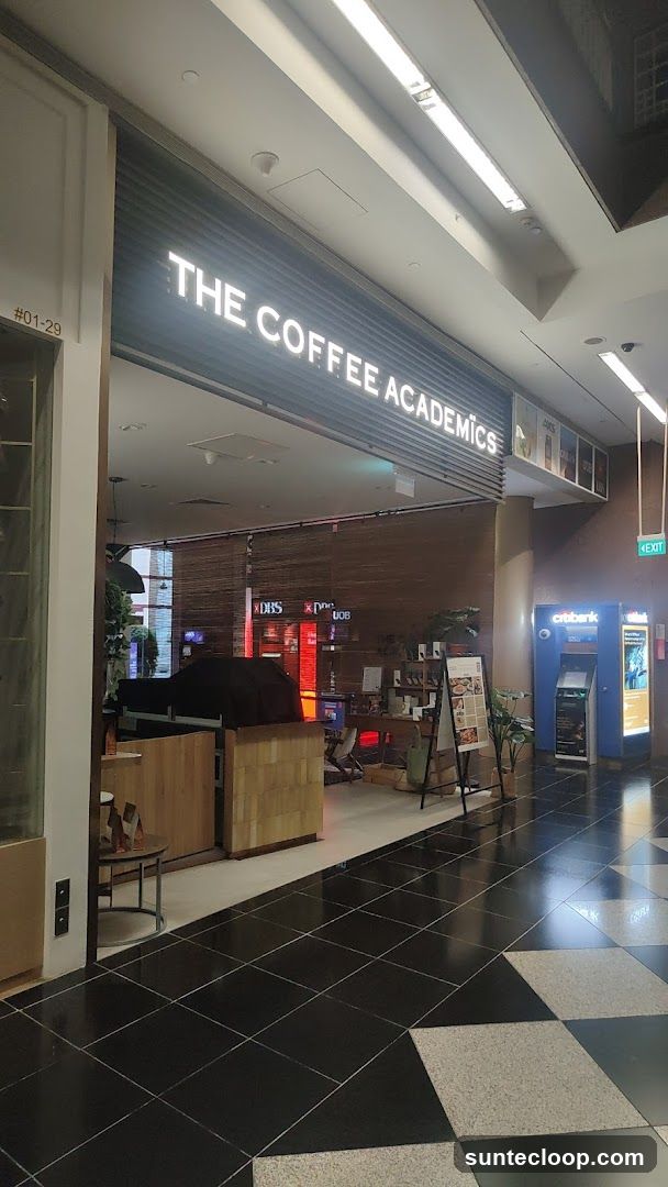 The exterior of a coffee shop with a prominent sign that reads "THE COFFEE ACADEMICS". The entrance is open, revealing a glimpse of the interior with wooden accents and some seating. To the right of the shop, there are ATMs and an exit sign.