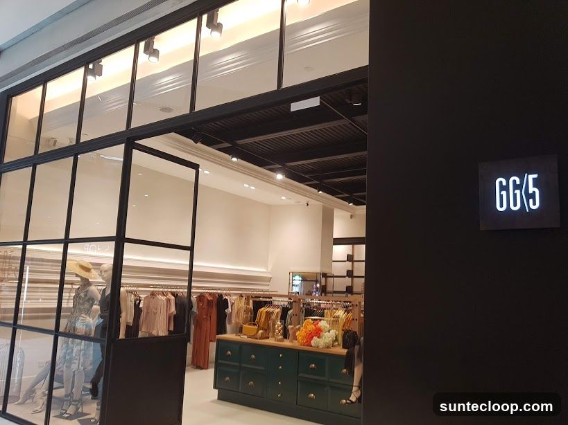 A view of a store entrance with a black facade and a lit sign that reads "GG/5". Through the glass windows, racks of clothing and mannequins are visible. A green display cabinet with drawers is also present.