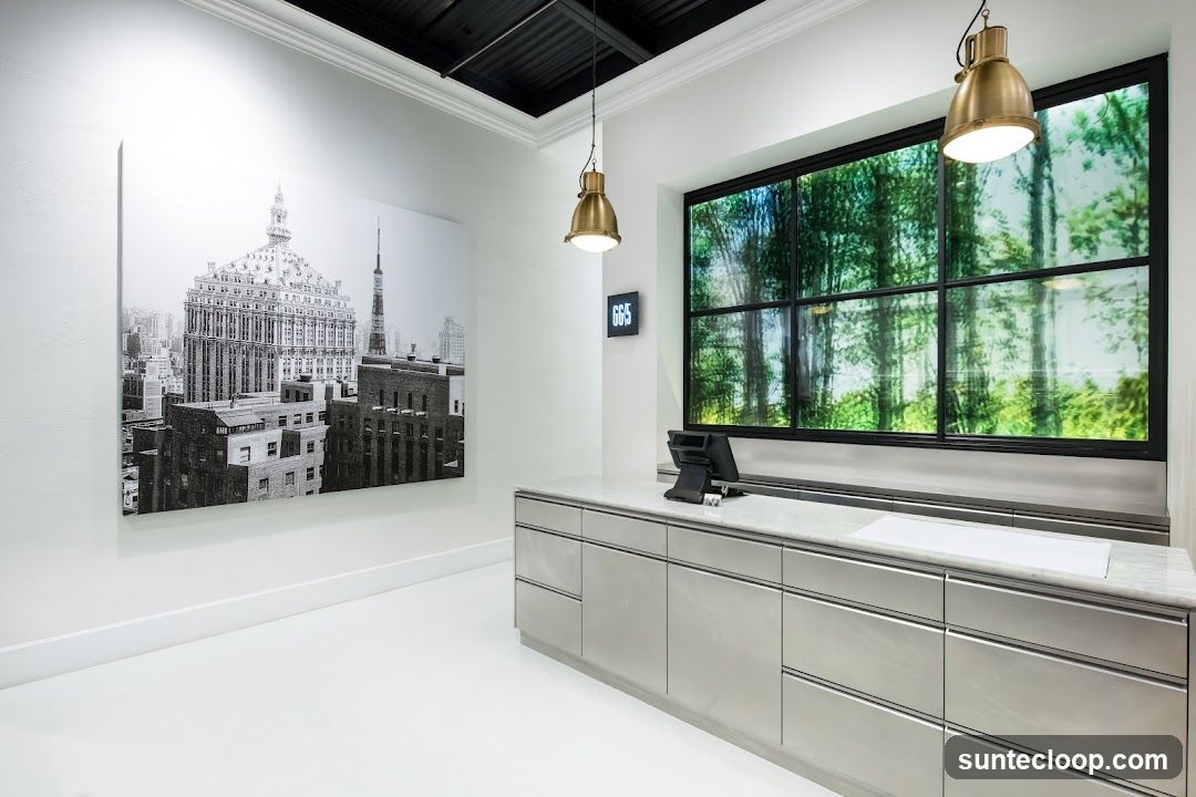 A sleek, modern interior space featuring a polished white floor and white walls. On the left, a large black and white canvas depicts a cityscape with a prominent ornate building. To the right, a counter with a marble top is equipped with a point-of-sale system. Above the counter, a large window is covered with a translucent film displaying a vibrant forest scene. Two industrial-style pendant lights hang from the black ceiling. A small digital display on the wall shows '665'.