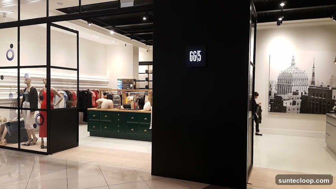 The image shows the interior of a clothing store. In the foreground, a black pillar partially obscures the view. To the left, a display window features mannequins dressed in fashionable attire, with clothing racks visible behind them. The store's interior is well-lit, showcasing a variety of garments on display. A large black and white mural of a cityscape adorns one of the walls. A person is seen walking in the background. A sign with the text "GG/5" is illuminated on a dark wall.