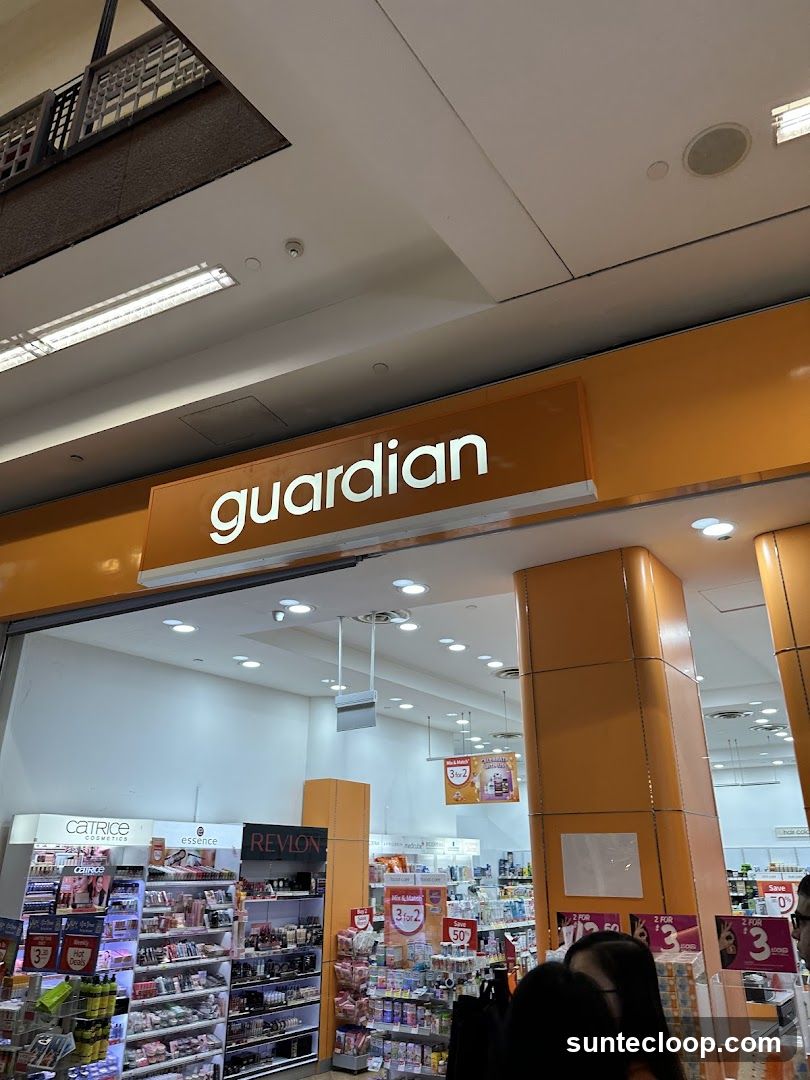 The exterior of a Guardian store, a health and beauty retailer. The prominent orange signage above the entrance reads "guardian" in white lettering. Inside, shelves are stocked with various cosmetics and personal care products from brands like Catrice, Essence, and Revlon. Promotional signs for "Mix & Match 3 for 2" and "Save 50%" are visible.