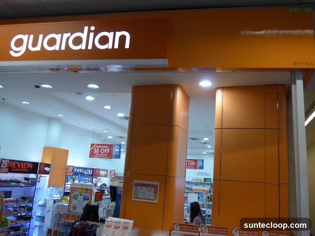 The exterior of a Guardian pharmacy with its prominent illuminated "guardian" logo above the entrance. Inside, shelves stocked with various health and beauty products are visible, along with promotional signs advertising discounts such as "ADDITIONAL $5 OFF" and "25% OFF REVLON".