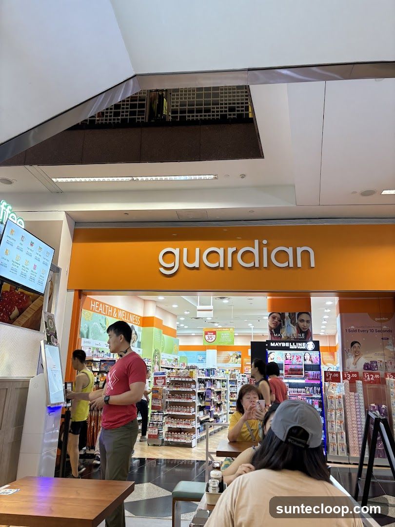The exterior of a Guardian store with its prominent orange signage displaying the store name. Inside, shelves stocked with health and wellness products are visible. Several customers are browsing or seated at tables. A digital menu board is also present to the left of the entrance.