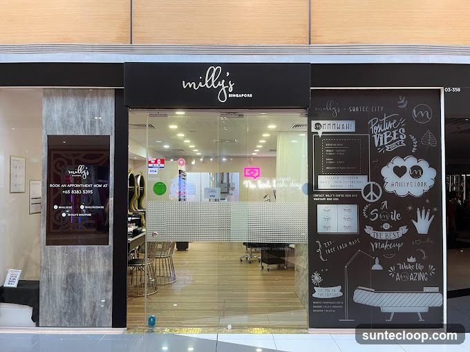 The exterior of a beauty salon called Milly's in Singapore. The storefront features a large glass window with frosted designs and text. Above the window is the salon's name in white script on a black background. To the left, a digital display shows the salon's name and contact information. The interior of the salon is visible through the window, showing chairs and equipment.
