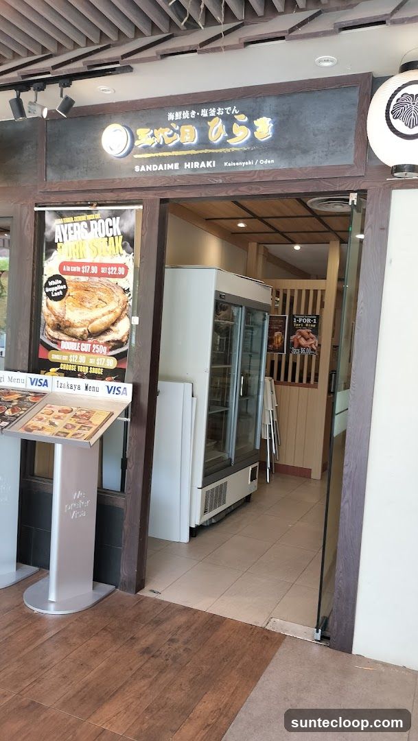 The exterior of a Japanese restaurant called Sandaime Hiraki, specializing in Kaisenyaki and Oden. The entrance features a prominent sign with Japanese characters and the restaurant name. To the left, a poster advertises "Ayers Rock Pork Steak" with pricing. A menu stand displaying "Izakaya Menu" is also visible. Inside, a refrigerator and seating area can be seen through the open doorway.