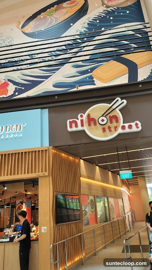 The exterior of a Japanese restaurant called Nihon Street. A large mural above the entrance depicts a bowl of ramen and sushi with waves. The restaurant has a modern wooden facade with a prominent sign. An exit sign is visible.