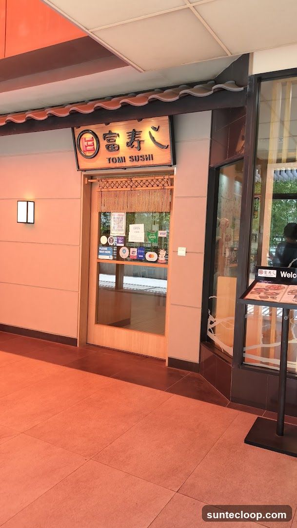 The exterior of a sushi restaurant with a wooden door and glass windows. A sign above the door reads "Tomi Sushi" in English and Japanese characters. Various payment method stickers and a "Please Wait Here" sign are visible on the door. A menu stand is placed to the right of the entrance.