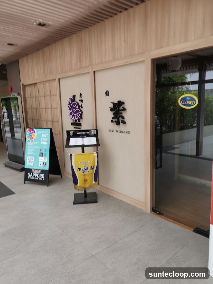 The exterior of a sushi restaurant with Japanese-style wooden paneling. A large sign with Japanese characters and "SUSHI MURASAKI" is visible on the wall. A "CLOSED" sign hangs on the glass door. Promotional signs for "galabunt" and "Sapporo Premium Beer" are placed outside. A banner for "Suntory Premium Malts" is also displayed.