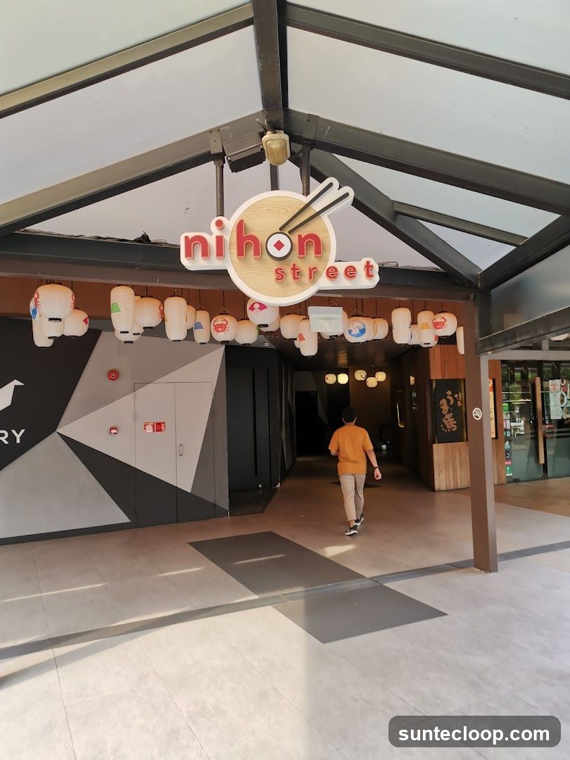 The exterior entrance of a Japanese restaurant called Nihon Street. A sign with the restaurant's name is visible above the entrance, featuring chopsticks and a red circle. Traditional Japanese lanterns hang in front of the entrance. A person is walking into the establishment.