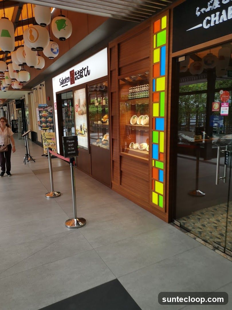 A row of Japanese restaurants with visible signage. The leftmost shop has a sign that reads 'Saboten' with Japanese characters. It features a display window showcasing food items and products. Next to it is another establishment with a wooden facade and a colorful stained-glass-like panel. To the right, a glass door leads into a restaurant with a black sign above it. A woman walks past the storefronts.