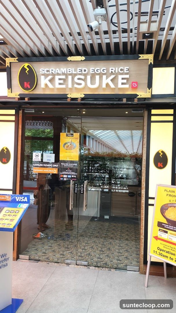 The exterior of a Keisuke Scrambled Egg Rice restaurant. The signage above the entrance reads "SCRAMBLED EGG RICE KEISUKE" with a stylized egg logo. The entrance has glass doors with various signs indicating payment methods and operating hours. A promotional stand with a picture of a dish is visible to the right.