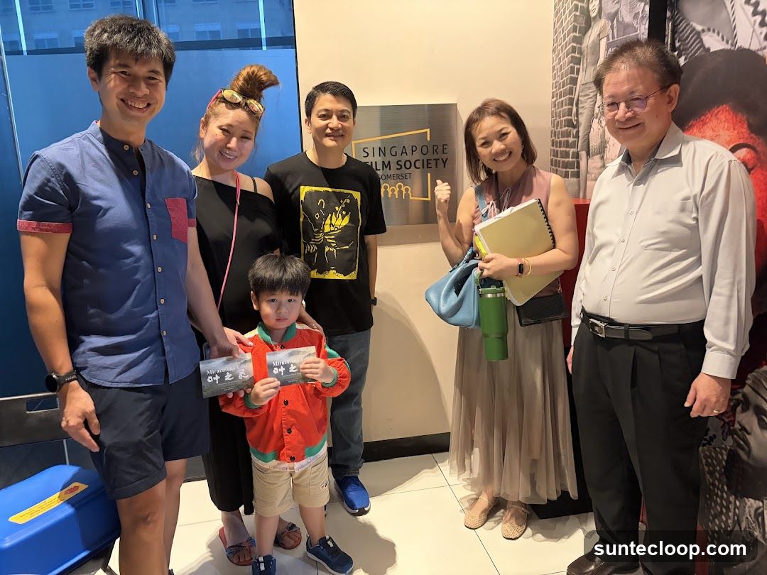 A group of six people, including a child, are posing for a photo in front of a sign that reads "SINGAPORE FILM SOCIETY SOMERSET". The child is holding two tickets. The people are smiling and appear to be attending an event.