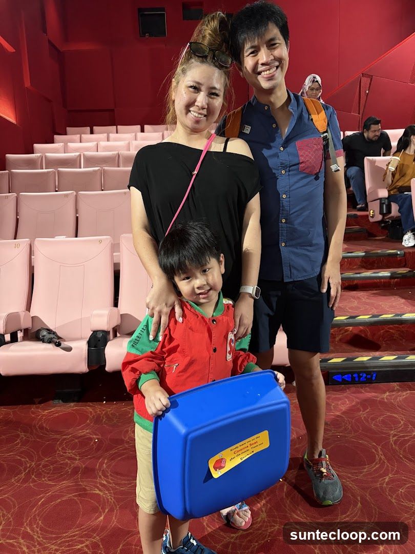 A family of three, including a young boy, are posing for a photo in a cinema. The boy is holding a blue booster seat. The seats in the cinema are pink and arranged in rows. The walls are red.