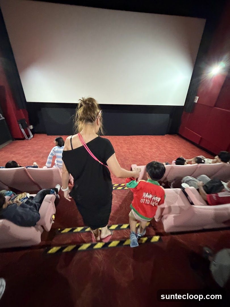 A woman and a child are walking down the aisle of a movie theater towards the screen. Other moviegoers are seated in the plush red seats. The screen is blank.