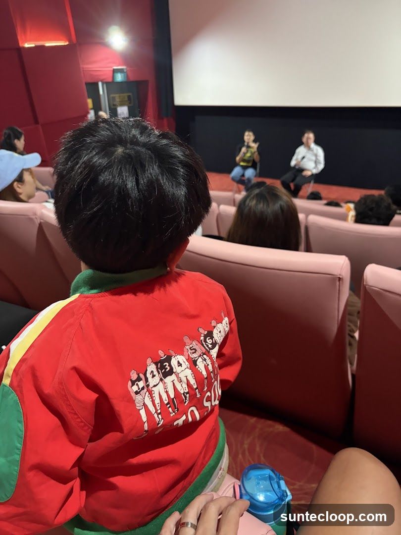 A child wearing a red jacket with a graphic of athletes on the back sits in a movie theater, facing a screen where two people are seated and speaking. The theater has pink seats and red walls.