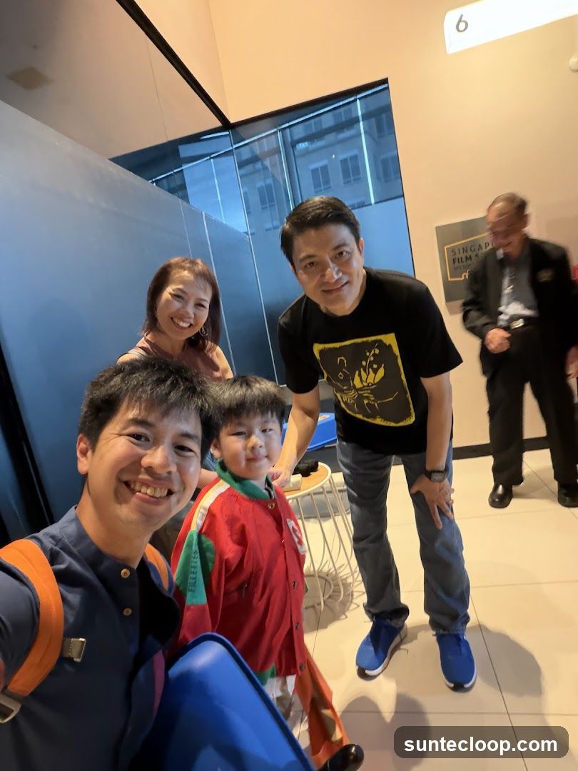 A group of four people, two adults and two children, are posing for a selfie in what appears to be a modern building. The adults are smiling at the camera, and the children are also looking towards the camera. In the background, a sign that reads "SINGAPORE FILM SOCIETY" is visible, along with a number "6" on the wall. The overall atmosphere seems casual and happy.