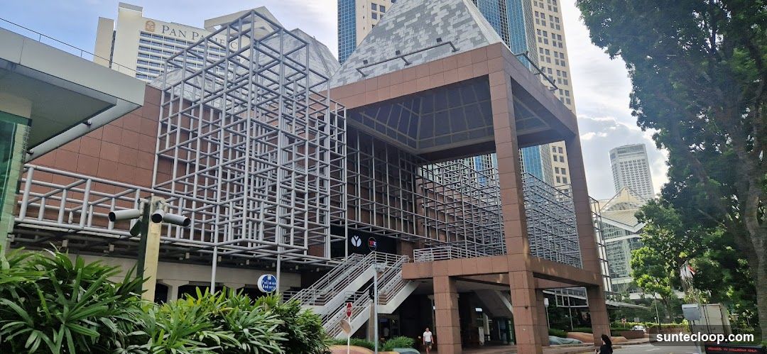 A wide shot of a modern building with a distinctive geometric metal lattice structure covering parts of its facade. The building has brown tiled walls and a pyramidal roof. In the background, several tall skyscrapers are visible, including the Pan Pacific hotel. Green trees and foliage are present in the foreground and to the right of the building. A few people are seen walking near the entrance.