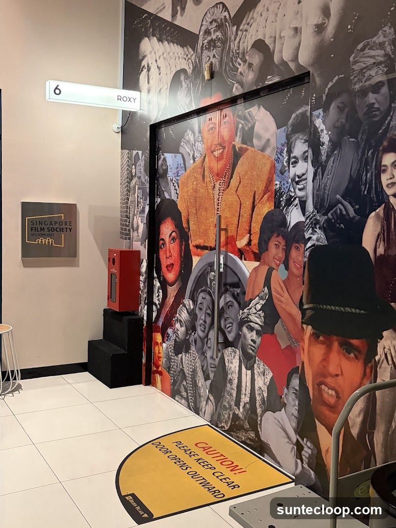 A doorway decorated with a collage of vintage film stills. A sign above the door reads "6 ROXY". To the left, a sign for "SINGAPORE FILM SOCIETY SFS SOMERSET" is mounted on the wall. A yellow mat on the floor reads "CAUTION! PLEASE KEEP CLEAR DOOR OPENS OUTWARD".