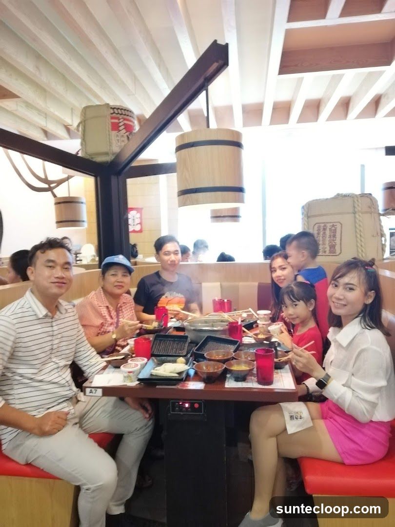 A group of people, including adults and children, are seated around a table in a restaurant. They are engaged in eating, with various dishes and utensils on the table. The restaurant appears to have a Japanese-inspired decor, with wooden elements and decorative barrels.