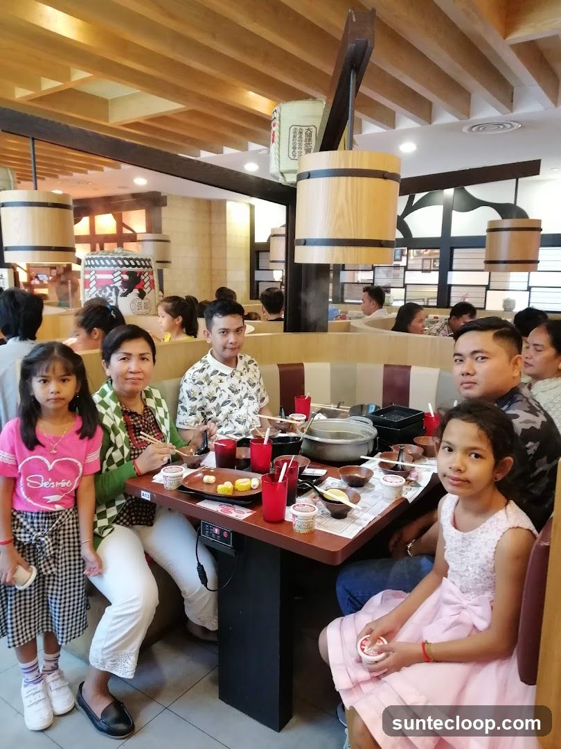 A family is seated around a table at a restaurant, with a hot pot in the center. The table is laden with food, drinks, and utensils. The restaurant has a modern interior with wooden accents and hanging lanterns. Several other diners are visible in the background.